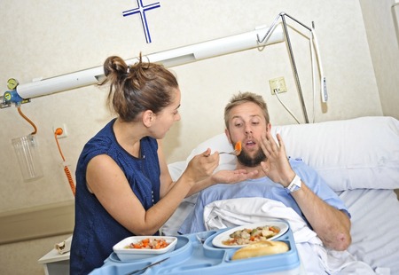 young wife trying to feed his husband lying in bed at hospital room ill after suffering accident and him looking unhappy with the diet food at the clinic center rejecting the mealの写真素材