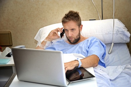young workaholic business man in hospital room lying in bed sick and injured working with mobile phone and computer laptop in health care and work obsession conceptの写真素材