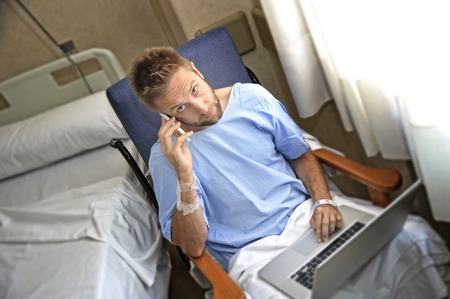 young workaholic business man in hospital room sick and injured after accident working with mobile phone and computer laptop in health care and work obsession conceptの写真素材