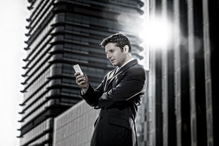 young attractive businessman in suit and necktie looking text message at mobile phone smiling happy standing outdoors in exterior in business district with lens flareの写真素材
