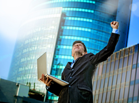 young attractive and successful businessman in suit and tie with computer laptop happy and excited doing victory sign celebrating success outdoor on financial district streetの写真素材