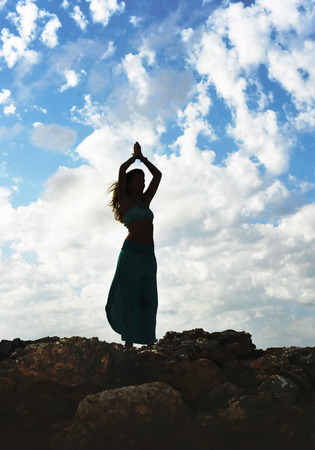 Silhouette of young attractive woman with dress and arms in Zen yoga pose at rock mountain looking at horizon under blue sky with clouds in relax and spiritual serenity and freedom conceptの写真素材