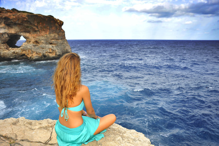 young attractive woman looking at sea horizon sitting alone on rock cliff in cyan glamour dress under a summer blue sky in relax , meditation and serenity conceptの写真素材