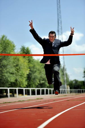 businessman running in suit and tie celebrating race victory on athletic track doing winning sign with arms in work and career success conceptの写真素材