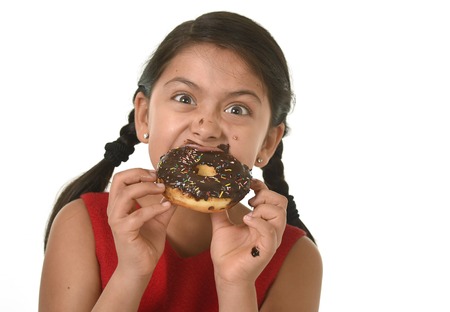hispanic female child in red dress eating chocolate donut with hands and mouth stained and dirty smiling happy in kid loving sugar and sweet food conceptの写真素材