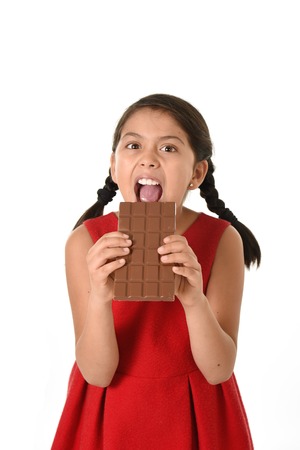 7 or 8 years old Hispanic girl wearing red dress holding with both hands big chocolate bar eating in happy and excited face expression in sugary nutrition and kids loving sweet conceptの写真素材