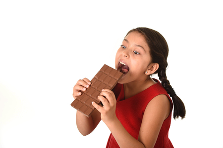 7 or 8 years old Hispanic girl wearing red dress holding with both hands big biting chocolate bar eating in happy and excited face expression in sugary nutrition and kids loving sweet conceptの写真素材