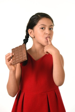 7 or 8 years old Hispanic girl wearing red dress holding big chocolate bar eating in happy and excited face expression licking her fingers in sugary nutrition and kids loving sweet conceptの写真素材