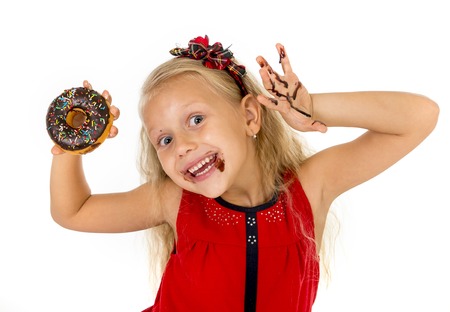 beautiful female child with blue eyes and long blond hair in cute red dress eating chocolate donut with syrup stains in mouth and dirty hand smiling happy isolated on white backgroundの写真素材