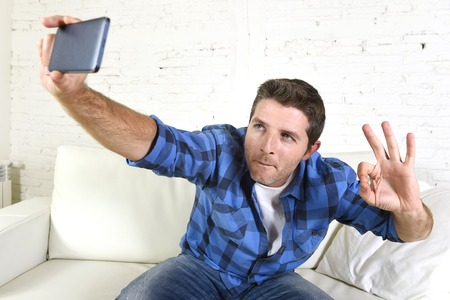 young attractive 30s man taking selfie photo or self video with mobile phone at home sitting on couch smiling happy giving thumbs up in use of technology and image conceptの写真素材