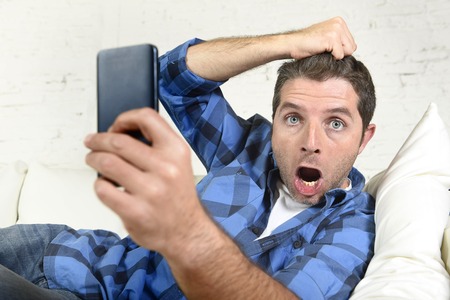 young attractive man lying at home couch using internet on his mobile phone looking surprised and shocked pulling hair in technology stress conceptの写真素材