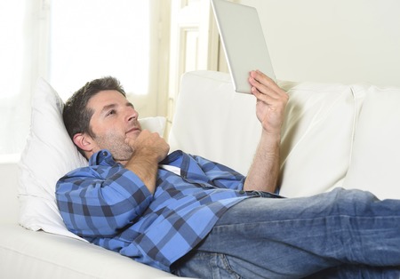 young attractive 30s man using digital tablet pad lying on couch at home living room networking looking relaxed and happy in portable technology and internet conceptの写真素材