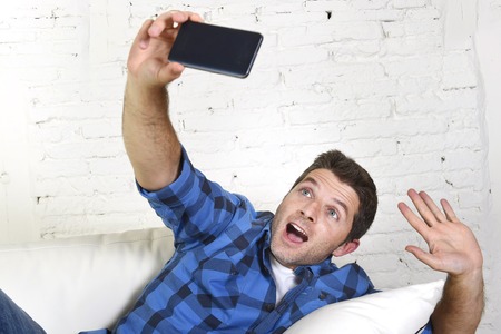 young attractive 30s man taking selfie picture or self video with mobile phone at home sitting on couch smiling happy in use of technology and image conceptの写真素材