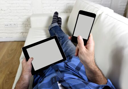 man lying on couch at home living room using simultaneously mobile phone and digital tablet pad in internet technology and social network addiction conceptの写真素材