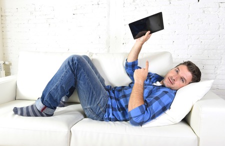 young attractive 30s man using digital tablet pad lying on couch at home living room networking looking relaxed and happy in portable technology and internet conceptの写真素材