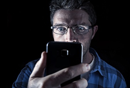 close up portrait of young man looking intensively to mobile phone screen with blue eyes wide open isolated on black background on dark edgy lighting scheme in addiction to internet technologyの写真素材