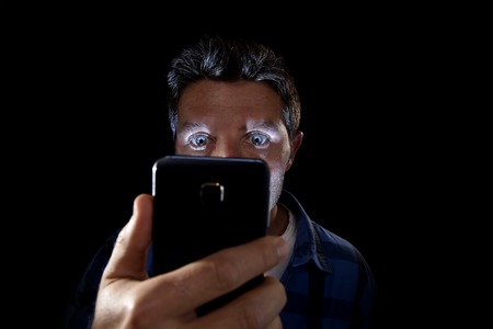 close up portrait of young man looking intensively to mobile phone screen with blue eyes wide open isolated on black background on dark edgy lighting scheme in addiction to internet technologyの写真素材