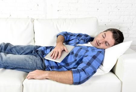 young attractive man in shirt and jeans sleeping at home white couch in living room having a rest after working with digital tablet pad in internet technology addiction and lifestyle conceptの写真素材
