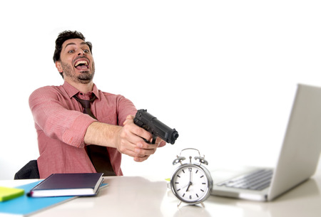young attractive businessman in stress at office computer desk pointing hand gun to alarm clock in out of time and project deadline expiring concept in angry  and frustreated face expressionの写真素材