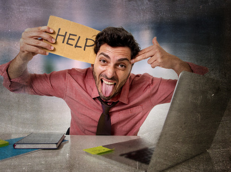 young businessman at office desk working on computer laptop asking for help holding cardboard sign looking sad and depressed making hand gun suicide sign in business stress and overwork conceptの写真素材