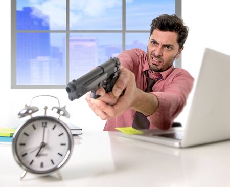 young attractive businessman working at computer desk pointing hand gun to alarm clock in out of time and business project deadline expiring concept angry and frustrated in business district officeの写真素材