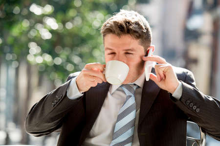 young attractive businessman sitting outdoors in coffee break holding cup and drinking while talking on mobile phone relaxed in communication and business conceptの写真素材