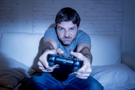 young excited man at home sitting on living room sofa playing video games using remote control joystick with freak intense face expression having fun in gaming addictionの写真素材