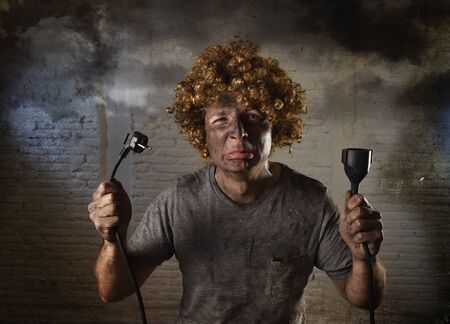 young man with funny curly wig holding electrical cable smoking after domestic accident with dirty burnt face and shock electrocuted expression in electricity DIY repairs danger concept in black smoke backgroundの写真素材