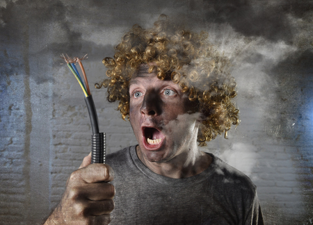 young man with funny curly wig holding electrical cable smoking after domestic accident with dirty burnt face and shock electrocuted expression in electricity DIY repairs danger concept in black smoke backgroundの写真素材
