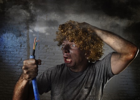 young man with funny curly wig holding electrical cable smoking after domestic accident with dirty burnt face and shock electrocuted expression in electricity DIY repairs danger concept in black smoke backgroundの写真素材