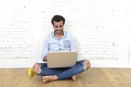 young attractive Latin man in hipster and modern casual style look sitting on unfurnished living room home floor working on laptop computer smiling happy in internet communication conceptの写真素材