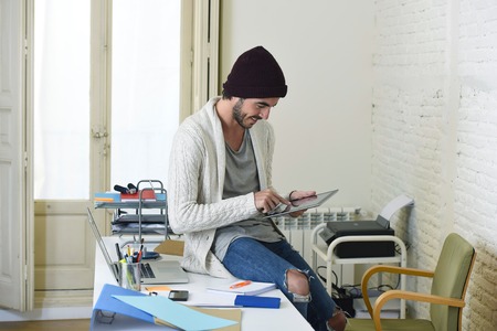 young trendy businessman in beanie and cool hipster informal look sitting on home office desk using internet on digital tablet pad happy and confident  in freelance business success conceptの写真素材