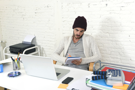 young trendy businessman in cool hipster beanie look writing on pad working in at modern home office with computer laptop as creative designer in freelance business conceptの写真素材