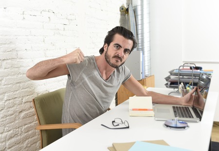young modern hipster style student or businessman working with laptop computer at home office looking angry upset and stressed gesturing with fist frustrated in business stress conceptの写真素材