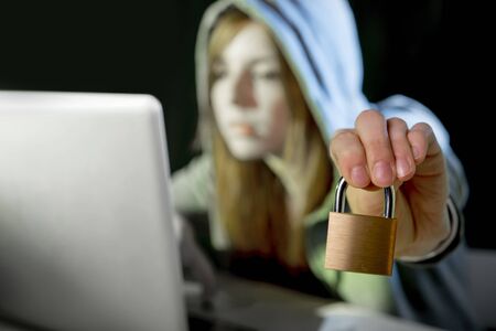 young attractive teen woman wearing hood on looking dark and dangerous hacking laptop computer system holding lock in cybercrime or cyber crime and internet criminal conceptの写真素材