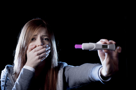 young sad woman scared and shocked holding pregnancy test positive result looking unhappy and in depression isolated on black background in unexpected pregnant woman conceptの写真素材