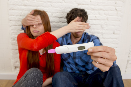 young sad couple in shock and surprise holding pink positive pregnancy test blocking eyes scared to confirm that the girl is pregnant  in unexpected baby and parenthood conceptの写真素材