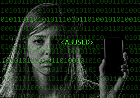 young scared and worried teenager girl holding mobile phone as internet stalked victim abused and cyberbullying or cyber bullying stress concept in binary code abused text black backgroundの写真素材