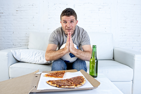 young man alone in stress watching football game on television sitting at home living room sofa couch with ball , pizza box and beer bottle enjoying the match praying nervous and excitedの写真素材