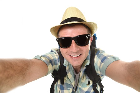 young attractive man or backpacker student taking selfie photo alone with mobile phone or camera wearing sunglasses and summer hat in tourist look smiling happy isolated on white backgroundの写真素材