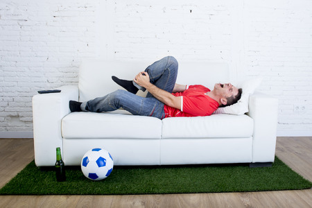 fanatic football fan lying on couch sofa with ball on green grass carpet emulating soccer stadium pitch mocking player in pain hurt on ankle in crazy supporter parody conceptの写真素材