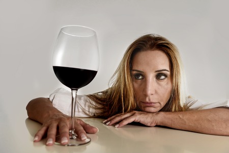 caucasian blond wasted and depressed alcoholic woman drinking red wine glass looking desperate and sad isolated on white in alcohol abuse and addiction and housewife alcoholism problemの写真素材