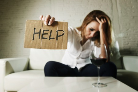 drunk alcoholic blond woman drinking whiskey glass asking for help holding message board depressed wasted and sad at home couch in alcohol abuse and housewife alcoholismの写真素材