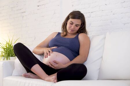 young beautiful and happy 8 or 9 months pregnant woman sitting at home living room couch holding big...