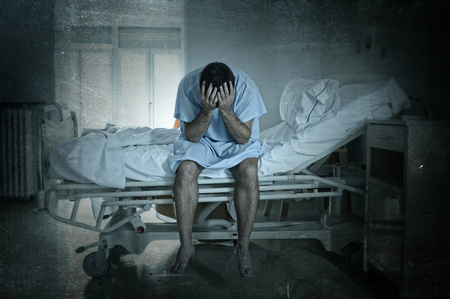 young desperate man sitting at hospital bed alone sad and devastated suffering depression crying at clinic for serious disease diagnose worried in fear on grunge dirty backgroundの写真素材