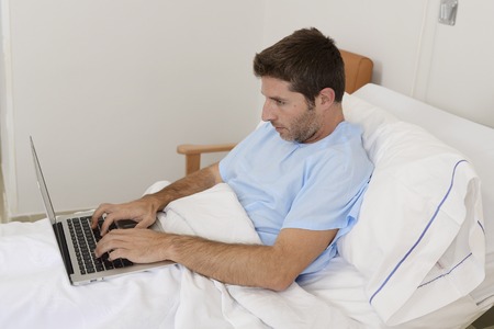 young businessman intern as patient in hospital suffering disease and working at the clinic bed with laptop computer in workaholic conceptの写真素材