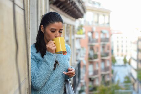 young beautiful latin woman drinking cup of coffee or tea holding the mug smiling happy at the apartment window balcony in relaxing breakfast at home conceptの写真素材