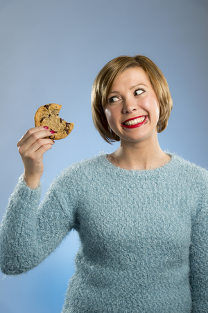 young blond cute beautiful caucasian woman with chocolate stain in her mouth eating big delicious cookie looking with temptation thinking if ignoring diet vs sweet junk food isolated backgroundの写真素材