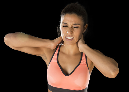 young attractive hispanic fitness woman touching and grabbing her neck and upper back suffering cervical pain isolated on black background in sport injury and body health care conceptの写真素材