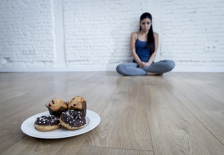 unhealthy sugar donuts and muffins and tempted young woman or teenager girl sitting on ground worried about overweight in diet and weight loss obsession in unhealthy nutrition conceptの写真素材